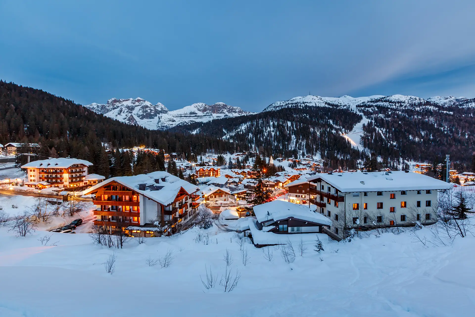 Veduta di un villaggio di montagna innevato al tramonto con luci accese e montagne sullo sfondo.