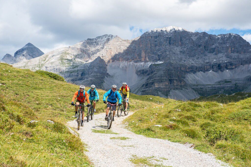 A group of cyclists rides on a mountain trail with rocky peaks in the background.