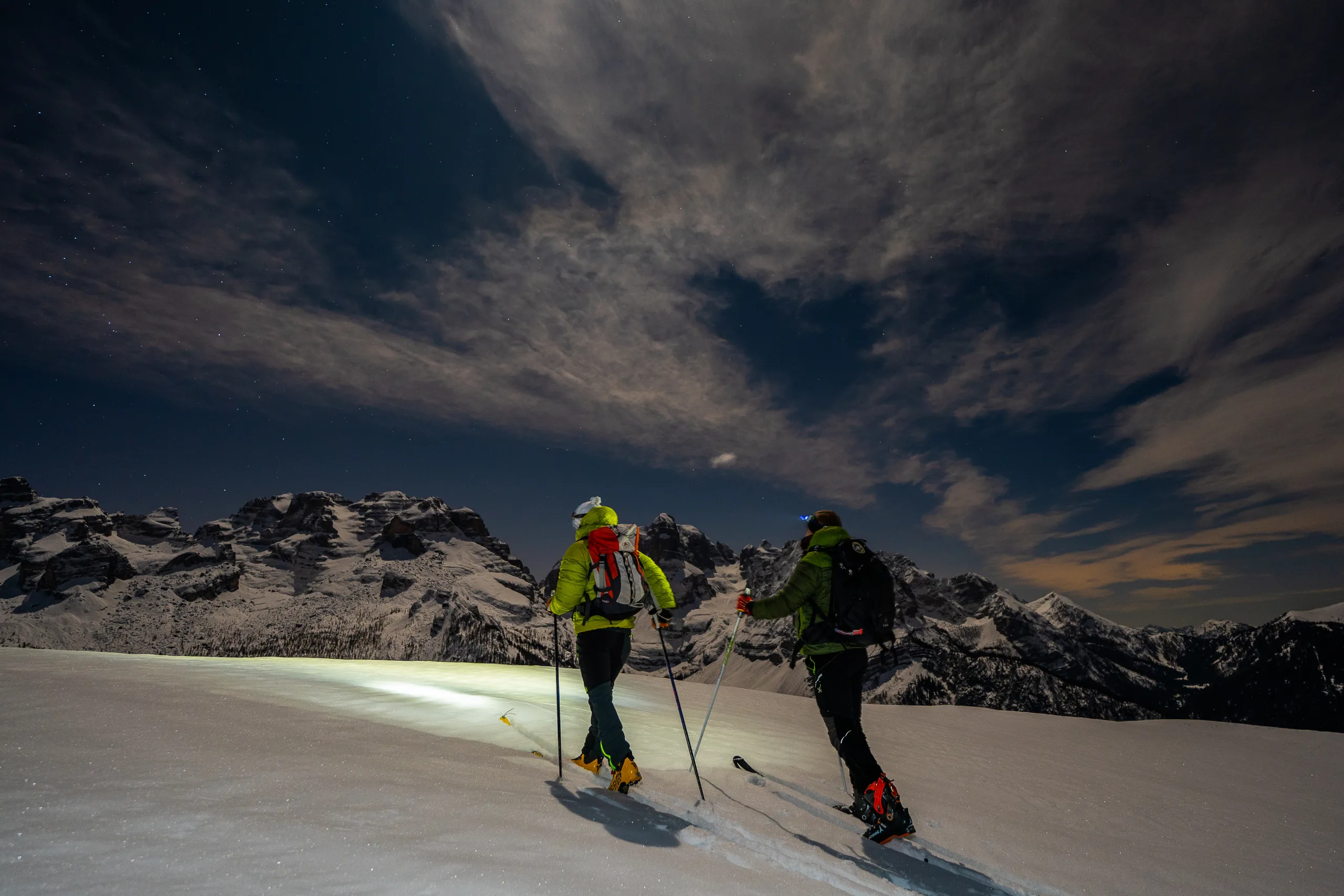 Due scialpinisti con frontale in salita di notte, con le Dolomiti di Brenta sullo sfondo