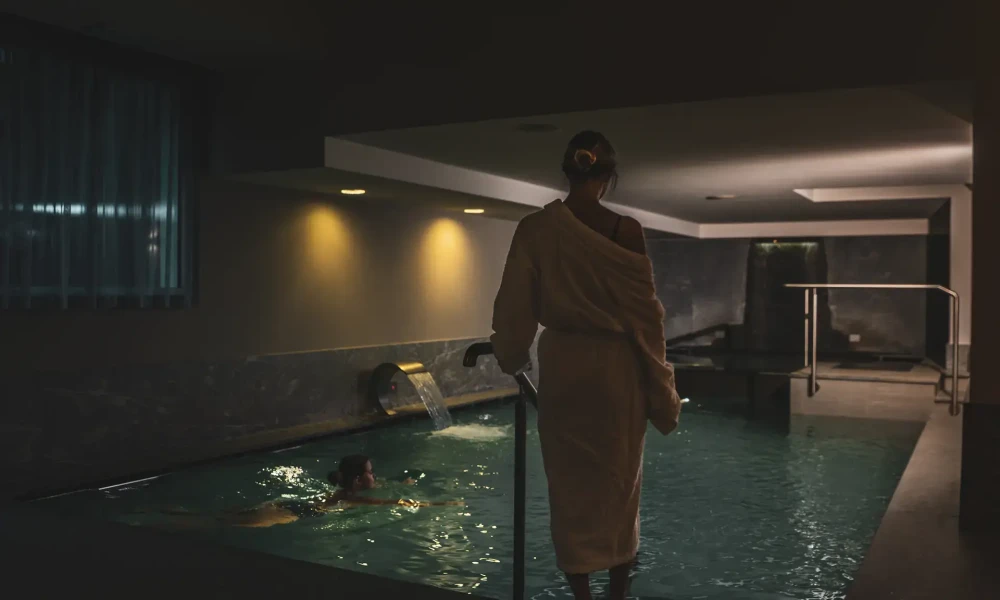 Persona in accappatoio osserva una piscina interna illuminata, con un'altra persona che nuota di sera.