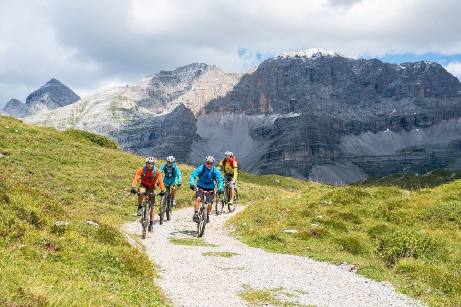 Cyclists riding on a mountain trail with rocky peaks in the background.