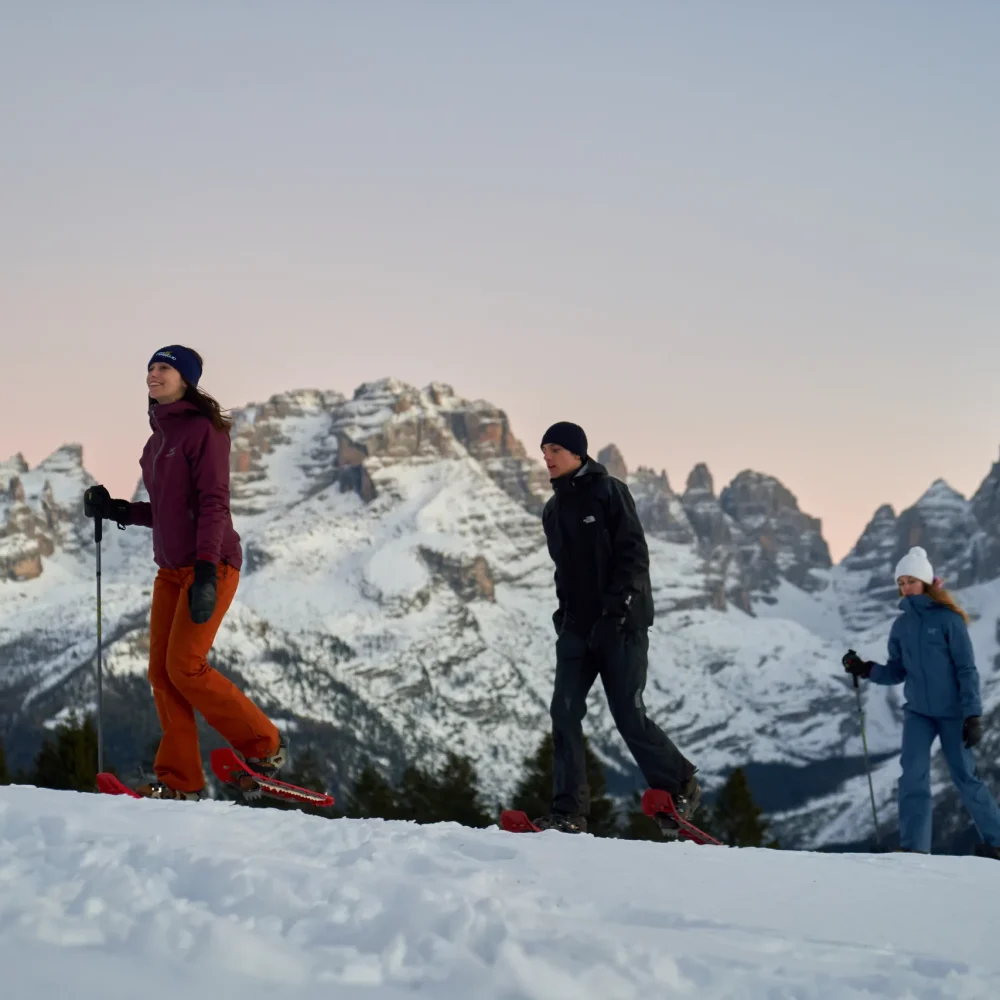 Tre persone con le ciaspole camminano sulla neve davanti alle Dolomiti di Brenta al tramonto