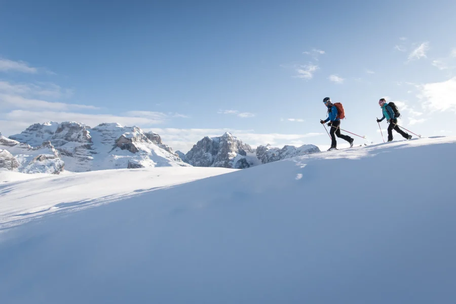 Due scialpinisti in salita su un pendio innevato, con le Dolomiti di Brenta sullo sfondo