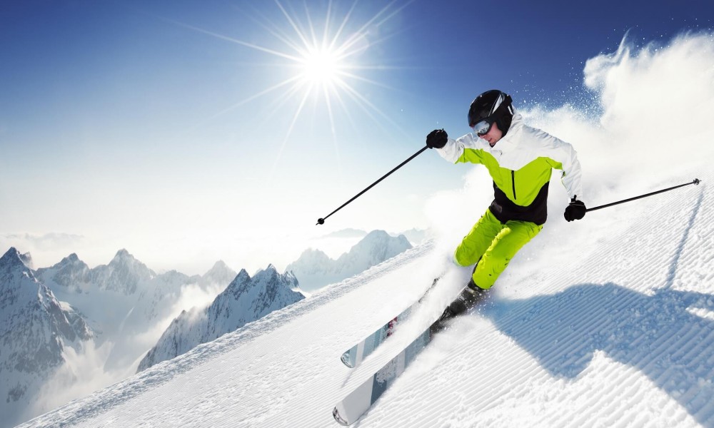 A skier in bright gear descends a snowy mountain under a clear blue sky.