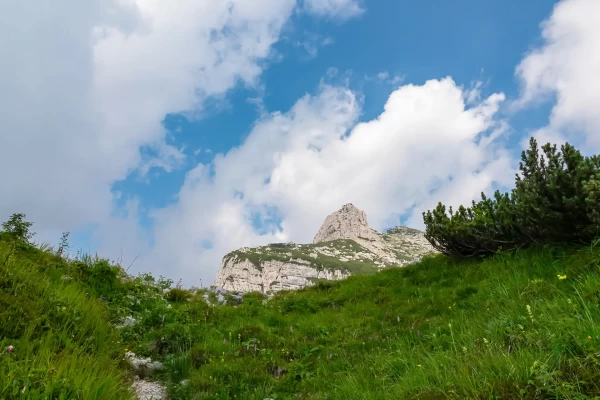 A rocky mountain peak under a blue sky with clouds, surrounded by green grassy slopes.